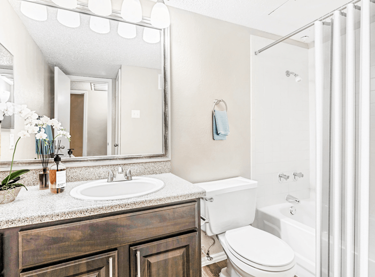 Virtually staged bathroom with wood cabinetry, granite style countertop, custom vanity mirror, white tile bathtub with sheer shower curtain